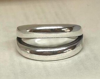 May include: A silver ring with a unique double-band design. The ring features two parallel, rounded bands connected by a small space, creating a modern and minimalist aesthetic. The polished metal reflects light, highlighting the smooth curves of the ring.