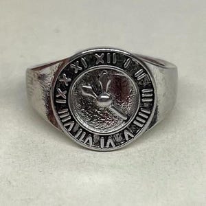Könnte beinhalten: Ein silberner Ring mit einem Zifferblatt-Design. Das Zifferblatt hat römische Ziffern und Zeiger, die eine bestimmte Uhrzeit anzeigen. Der Ring besteht aus glänzendem Metall.