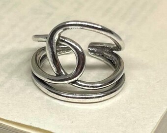 May include: A silver-toned ring with a unique, intertwined design. The ring features three bands that loop and cross over each other, creating an open, adjustable style. The metal has a slightly aged appearance, adding to its character.