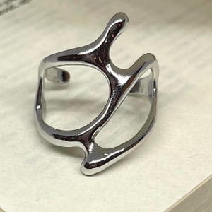 May include: A silver-colored ring with an abstract, organic design. The ring features a fluid, openwork structure with curved lines and irregular shapes, creating a modern and artistic aesthetic. The ring is likely made of metal and has a polished, reflective surface.