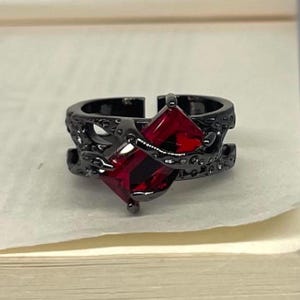 May include: A black ring featuring a square, red gemstone. The ring has an intricate, textured pattern around the band and setting. The gemstone is a rich red, and the ring seems to be made of metal.