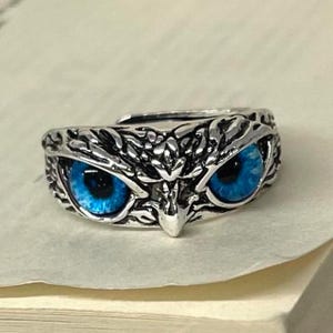May include: A silver-coloured ring shaped like an owl's face, with intricate feather detailing. The ring has vivid blue eyes that stand out against the metal. The adjustable band ensures a comfortable fit.