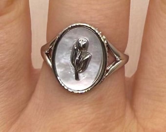 May include: A silver ring with an oval mother-of-pearl face. The face features a dark, stylized floral design. The ring is set on a finger, showcasing its intricate details and elegant design. The ring's band has a unique, open design.