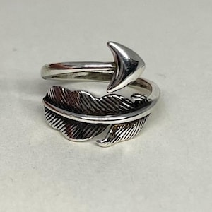 Könnte beinhalten: Silberfarbener Ring mit Feder- und Pfeildesign. Der Ring umschließt den Finger, mit einer detaillierten Feder am unteren Teil und einem nach oben zeigenden Pfeil. Der Ring ist wahrscheinlich aus Metall gefertigt.