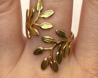 May include: A gold-colored ring featuring a leaf design. The ring wraps around the finger with two branches of leaves. The leaves have a smooth, polished finish. The ring is worn on a finger.