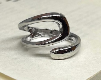 May include: A shiny silver ring with a unique, open design. The ring features a curved, abstract shape that wraps around the finger. The ring is displayed on a light-colored surface, highlighting its polished finish.