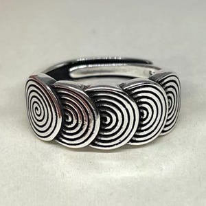 Peut inclure: Une bague en argent ornée d'une série de spirales reliées. La bague a un design ouvert, permettant un ajustement de la taille. Les spirales sont noires et blanches, créant un contraste saisissant. La bague est probablement en argent sterling.
