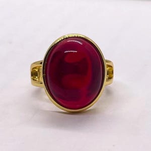 May include: A gold ring with a large, oval, deep red gemstone. The gemstone is set in a gold bezel, and the ring has a simple design. The ring is a piece of jewellery.