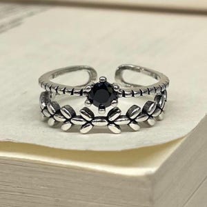 Op de afbeelding: Een verstelbare ring in zilverkleur met een zwarte edelsteen in het midden. De ring heeft een dubbelbandontwerp, met een bovenste band met kralen en een onderste band versierd met bladvormige details. De ring staat tegen een neutrale achtergrond.
