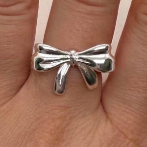 Può includere: Un anello color argento a forma di fiocco. Il fiocco presenta un design dettagliato con anelli curvi e un nodo centrale. L'anello è indossato su un dito, mettendo in risalto lo stile elegante e femminile del gioiello.