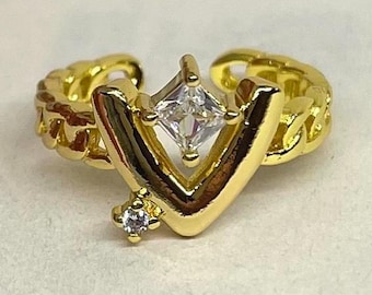 May include: A gold-colored ring with a V-shaped design and a square, clear gemstone at the center. The ring has a chain-like band and a small, round gemstone at the bottom of the V.