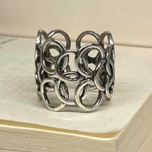 Peut inclure: Une bague argentée présentant un motif répétitif de cercles interconnectés. La bague a un design à large bande et un aspect texturé. Le bijou est présenté sur une surface de couleur claire, mettant en valeur ses détails complexes et son éclat métallique.