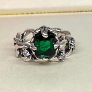Puede incluir: Un anillo de plata con una gran gema verde esmeralda de corte ovalado como pieza central. El anillo tiene un diseño detallado y ornamentado con gemas claras más pequeñas y un motivo floral.
