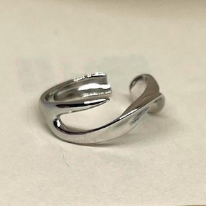 Pode incluir: Um anel prateado com um design aberto único. O anel apresenta uma banda lisa e curva que envolve o dedo, com um espaço aberto no topo. O anel é feito de um metal brilhante e refletor.
