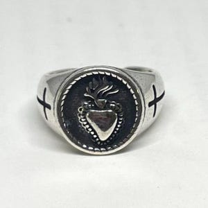 May include: Silver signet ring with a central heart and flame design, encircled by a beaded edge. Crosses are etched on the sides. The ring is a bold statement piece.