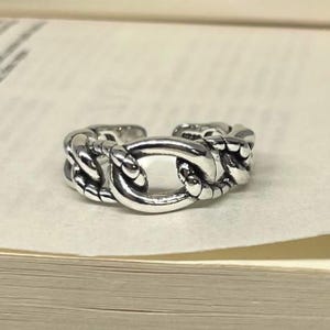 Peut inclure: Une bague argentée avec un motif de chaîne. La bague présente des maillons ovales et arrondis interconnectés, créant un aspect texturé. La bague est ouverte à l'arrière, permettant un ajustement de la taille. La bague est en métal brillant.