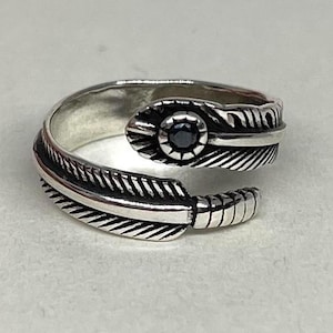 May include: A silver ring with a feather design. The ring has a central black gemstone and detailed feather engravings. The ring's design wraps around the finger, creating a unique and stylish accessory.