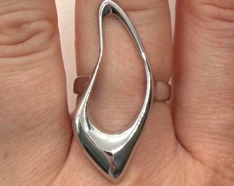 May include: A silver-colored ring with a unique, open teardrop design. The ring is smooth and reflective, with a polished finish. The ring is worn on a finger, showcasing its modern, abstract shape. The jewelry piece is a statement accessory.