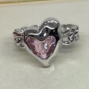 May include: A silver ring with a heart-shaped design. The heart has a pink gemstone centre and is surrounded by a chain-link band. The ring is a close-up shot, showcasing the details of the jewellery.