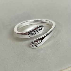 May include: A silver ring featuring the word "FAITH" engraved on the top. The ring has a cross design on the end. The ring is a wrap-around style.