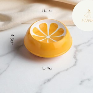 May include: A bright orange ceramic bowl with a white citrus fruit design on the inside. The bowl is 1.5 inches in diameter and 1.7 inches tall. The bowl is on a white marble surface.