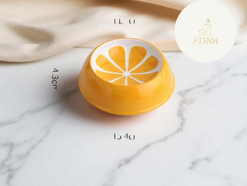 May include: A bright orange ceramic bowl with a white citrus fruit design on the inside. The bowl is 1.5 inches in diameter and 1.7 inches tall. The bowl is on a white marble surface.