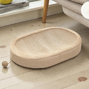 Puede incluir: Rascador para gatos beige, de forma ovalada y textura tejida. Una pequeña bola redonda con una cuerda está unida al lateral. El rascador está sobre un suelo de madera clara, cerca de un sillón beige.