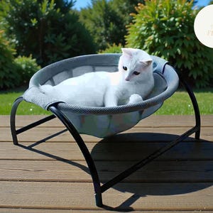 May include: A white cat lounges in a grey and black pet bed. The cat bed features a mesh fabric and is supported by a black metal frame. The cat is resting on a wooden deck, with green foliage in the background.