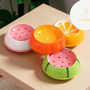 May include: Four ceramic pet bowls with fruit designs. The bowls are shaped like a watermelon, orange, and strawberry. The watermelon bowl is green with black seeds, the orange bowl is orange with a slice design, and the strawberry bowl is pink with red seeds.