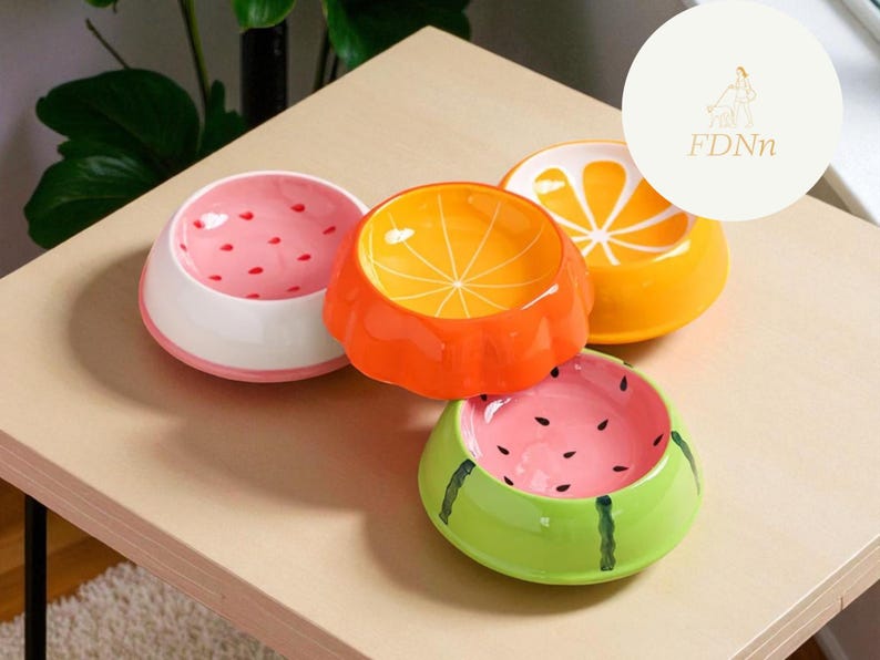 May include: Four ceramic pet bowls with fruit designs. The bowls are shaped like a watermelon, orange, and strawberry. The watermelon bowl is green with black seeds, the orange bowl is orange with a slice design, and the strawberry bowl is pink with red seeds.