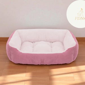 K&ouml;nnte beinhalten: Ein rosa rechteckiges Haustierbett mit einem Cord-Au&szlig;enmaterial und einem weichen, wei&szlig;en Innenraum. Das Bett steht auf einem Holzboden. Ein wei&szlig;er Kreis mit einem goldenen Logo und den Buchstaben "FDnn" befindet sich im Hintergrund.
