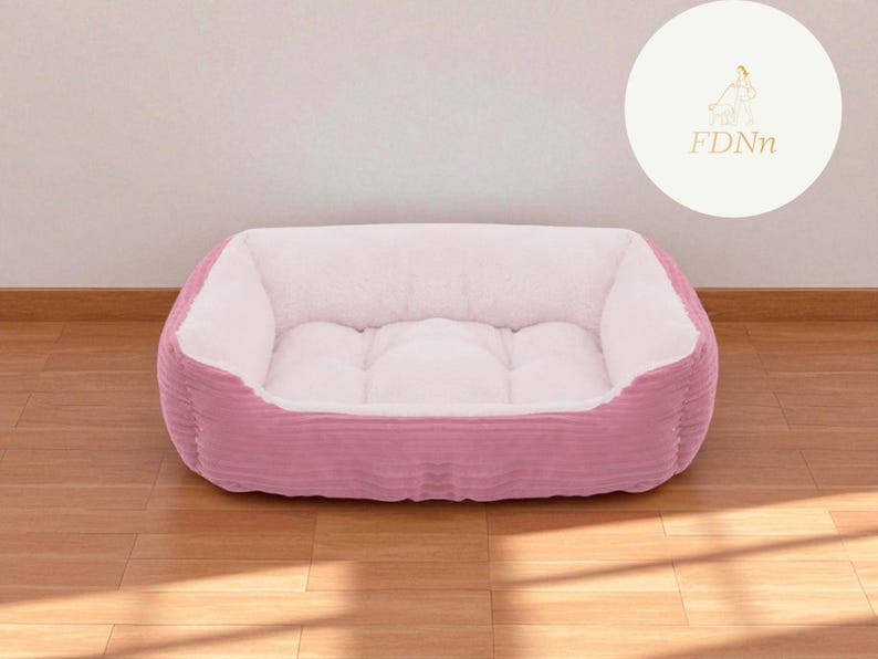 K&ouml;nnte beinhalten: Ein rosa rechteckiges Haustierbett mit einem Cord-Au&szlig;enmaterial und einem weichen, wei&szlig;en Innenraum. Das Bett steht auf einem Holzboden. Ein wei&szlig;er Kreis mit einem goldenen Logo und den Buchstaben "FDnn" befindet sich im Hintergrund.