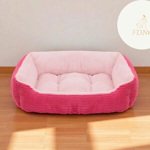 K&ouml;nnte beinhalten: Ein pinkfarbenes Haustierbett mit einem weichen, hellrosa Innenraum und einem Cord-Au&szlig;enmaterial. Das Bett hat eine rechteckige Form mit erh&ouml;hten Seiten und einer gepolsterten Mitte. F&uuml;r kleine Haustiere.