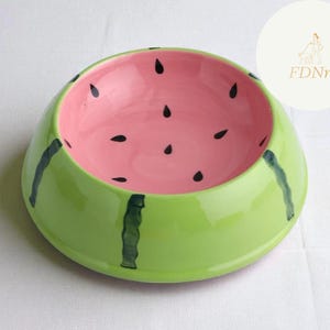 May include: A ceramic pet bowl in the shape of a watermelon. The bowl is painted with a bright green exterior, a pink interior, and black seed details. The bowl has dark green stripes on the exterior.