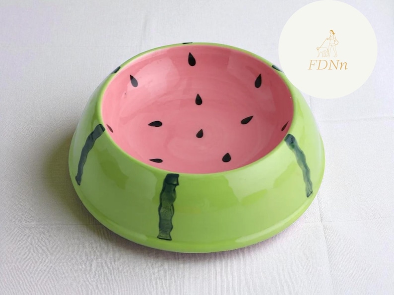 May include: A ceramic pet bowl in the shape of a watermelon. The bowl is painted with a bright green exterior, a pink interior, and black seed details. The bowl has dark green stripes on the exterior.