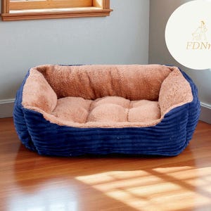 K&ouml;nnte beinhalten: Ein rechteckiges Haustierbett mit marineblauem Cord-Au&szlig;enmaterial und einem weichen, beigefarbenen Innenraum. Das Bett steht auf einem Holzboden. Das Design deutet auf einen komfortablen und warmen Platz f&uuml;r ein kleines Haustier hin.