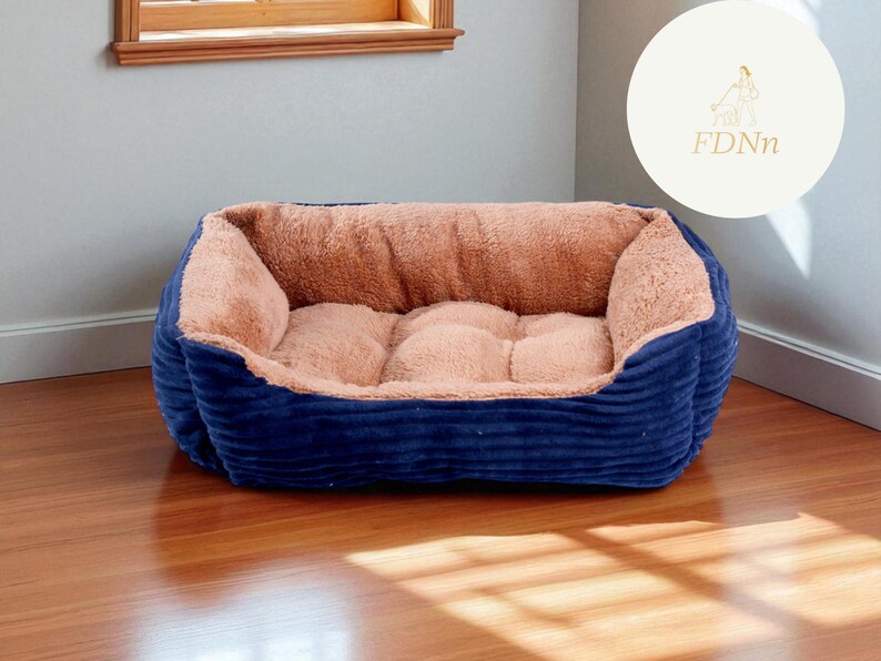 K&ouml;nnte beinhalten: Ein rechteckiges Haustierbett mit marineblauem Cord-Au&szlig;enmaterial und einem weichen, beigefarbenen Innenraum. Das Bett steht auf einem Holzboden. Das Design deutet auf einen komfortablen und warmen Platz f&uuml;r ein kleines Haustier hin.