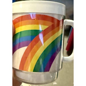 May include: A clear plastic mug with a white handle and rim. The mug features a rainbow design with red, orange, yellow, green, blue, and purple arches. The mug is designed to be stackable.