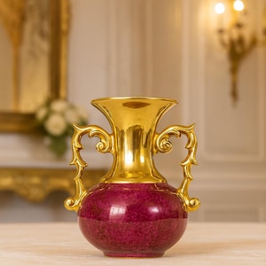 May include: A decorative vase with a gold-colored top and handles, and a deep red, speckled body. The vase has a classic urn shape, with ornate, curved handles. The gold top and handles provide a striking contrast to the red body.
