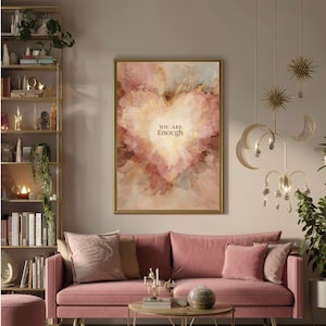 May include: A framed artwork with a gold border features a heart-shaped design in shades of pink, peach, and gold. The words "YOU ARE Enough" are centered within the heart. The artwork hangs above a pink sofa with matching pillows.