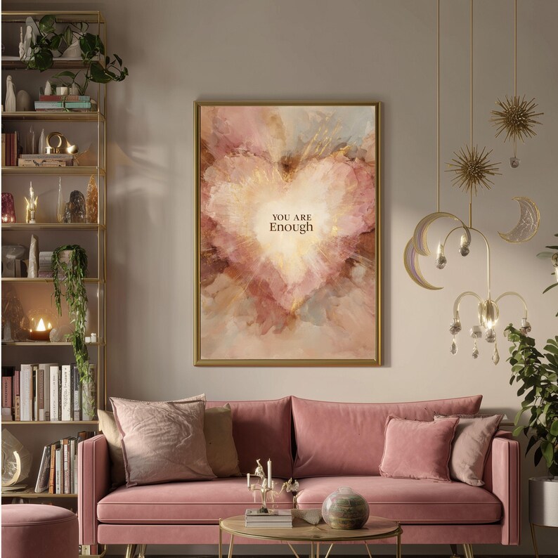 May include: A framed artwork with a gold border features a heart-shaped design in shades of pink, peach, and gold. The words "YOU ARE Enough" are centered within the heart. The artwork hangs above a pink sofa with matching pillows.