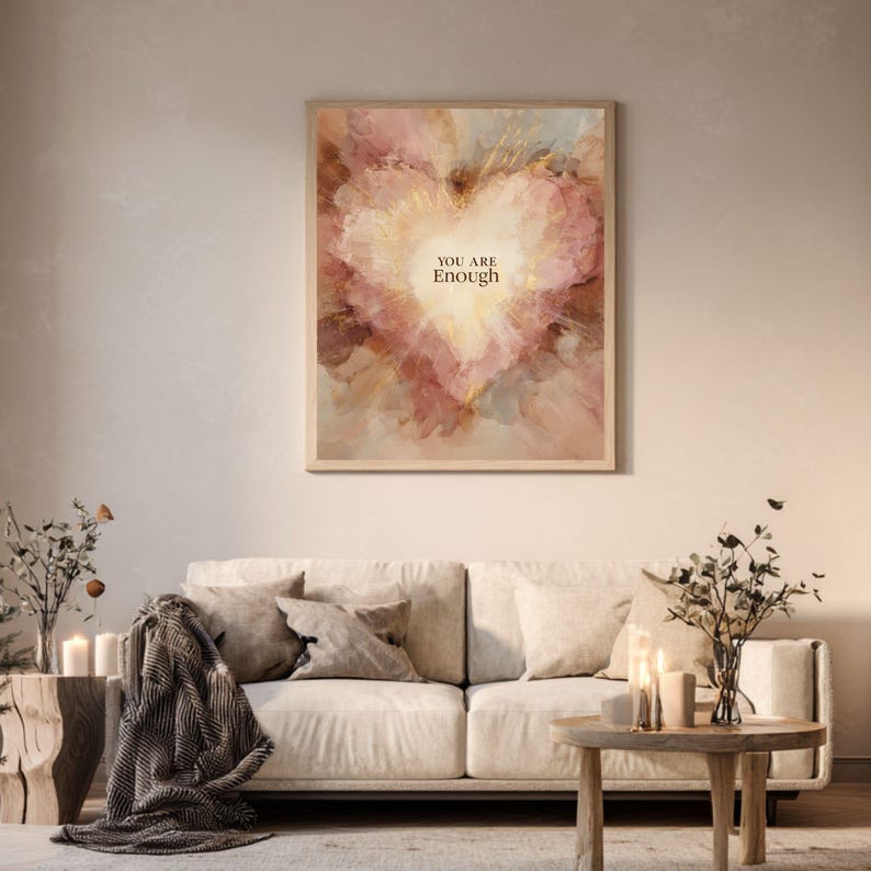 May include: A framed art print with a heart-shaped design in shades of pink, peach, and gold. The words "YOU ARE Enough" are centered within the heart. The artwork is displayed above a cream-colored sofa in a living room setting.