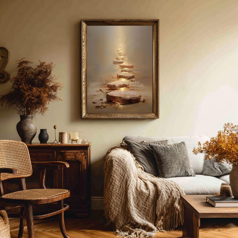 Op de afbeelding: Een ingelijst kunstwerk toont een pad van verlichte stapstenen dat naar een helder licht leidt. Het kunstwerk bevindt zich in een gouden lijst en hangt boven een bank met een geweven sprei. De kamer heeft een neutraal kleurenpalet met houten meubels.