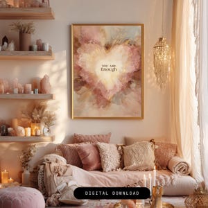 May include: A framed print with a heart-shaped design in shades of pink, gold, and beige, with the words "YOU ARE Enough" in the center. The artwork hangs above a sofa with pink and beige pillows. The room is decorated in a bohemian style.