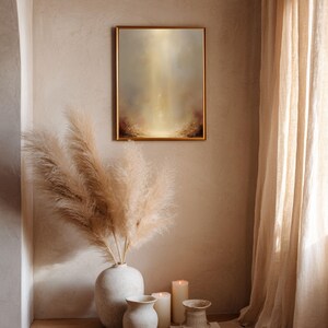 May include: A framed abstract painting with a gold frame hangs on a beige wall. The artwork features a soft, ethereal design with warm, golden light and hints of red and brown. A vase with tall, feathery pampas grass, candles, and other decorative vases are in the foreground.