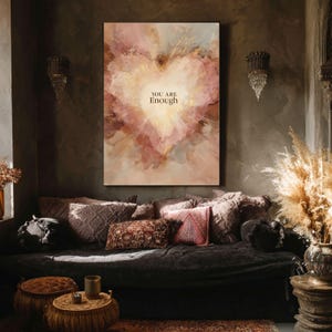 May include: A large canvas art piece featuring a heart-shaped design in shades of pink, gold, and cream. The words "YOU ARE Enough" are centered within the heart. The artwork hangs above a dark-colored sofa with decorative pillows.