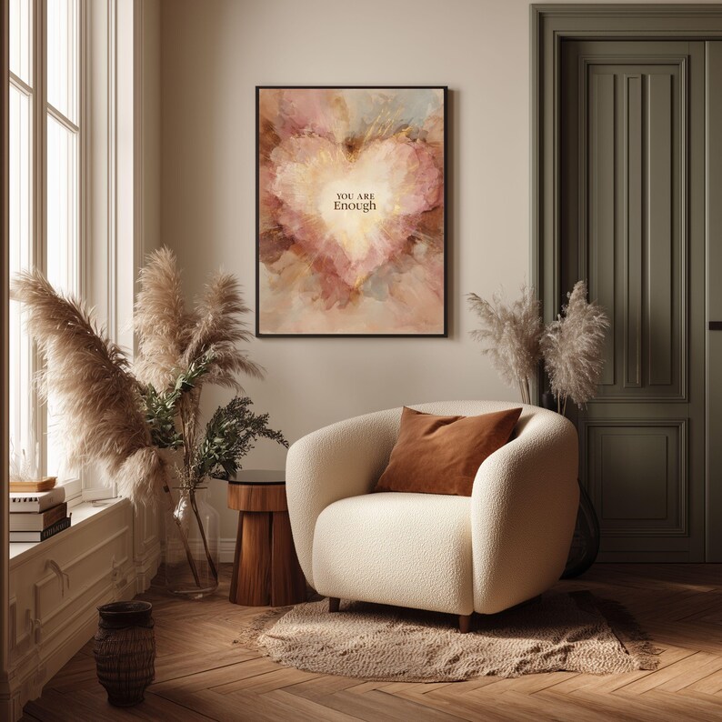 May include: A framed art print with a heart-shaped design in shades of pink, peach, and brown, with the words "YOU ARE ENOUGH". The artwork hangs above a cream-colored armchair with a brown pillow. The room has a neutral color palette.