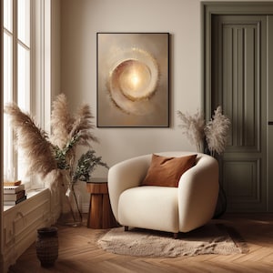 May include: A framed abstract art piece with a spiral design in beige and gold tones hangs above a cream-colored armchair. The room features a wooden floor, pampas grass, and a brown throw pillow.