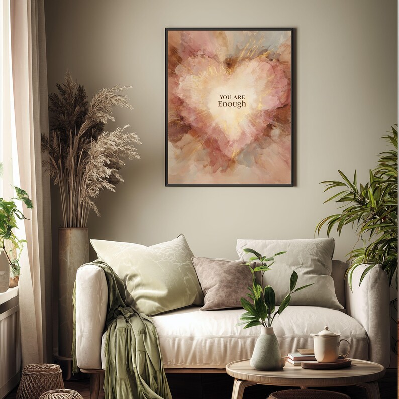 May include: A framed art print with a heart-shaped watercolor design in shades of pink, gold, and brown. The words "YOU ARE Enough" are centered within the heart. The print hangs above a cream-colored sofa with decorative pillows.