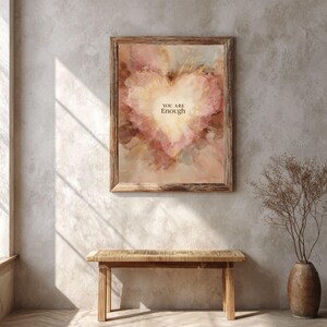 May include: A framed art print with a heart-shaped design in shades of pink, peach, and gold. The text "YOU ARE Enough" is centered within the heart. The print is displayed above a wooden bench.